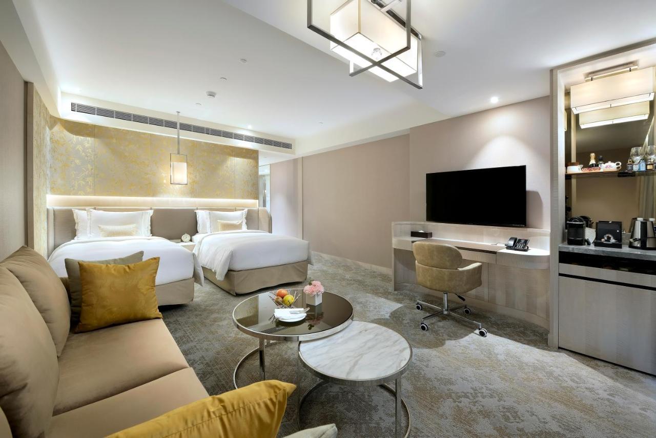 Caesar Park Hotel Banqiao Taipei Exterior photo The photo shows a luxurious hotel room featuring two beds with white linens, a comfortable seating area with a sofa and decorative pillows, and a coffee table. There is a large flat-screen TV mounted on the wall opposite the beds, along with a worksp